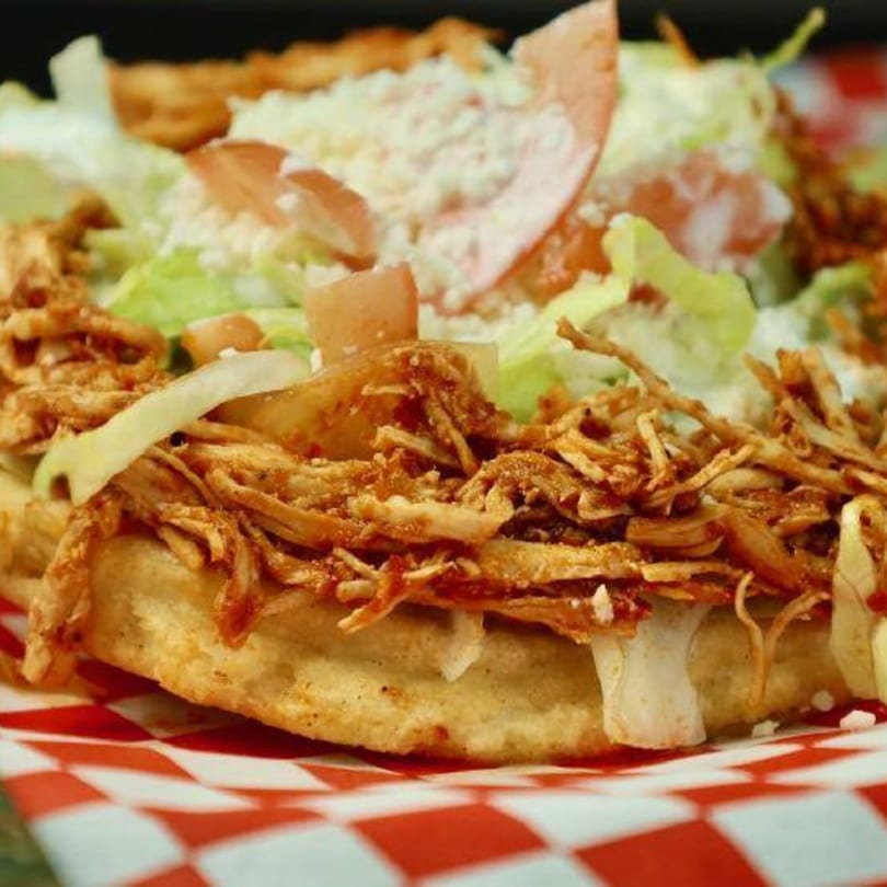 Close-up of a Mexican sope with shredded chicken, lettuce, tomato, and cheese on checkered paper.