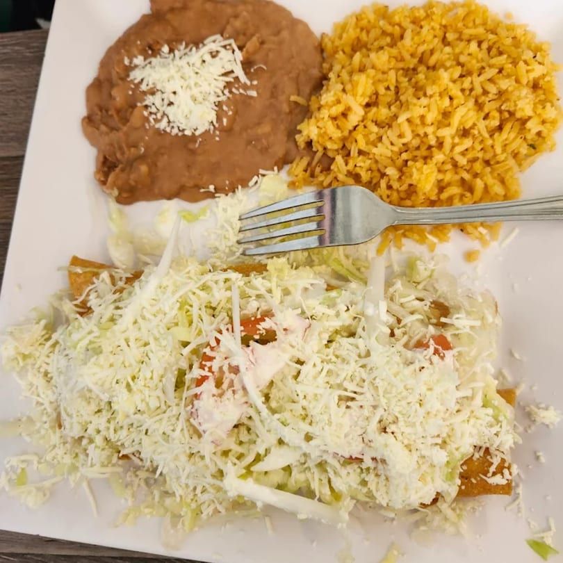 A plate of taquitos topped with shredded lettuce, cheese, and sour cream, with rice and beans.