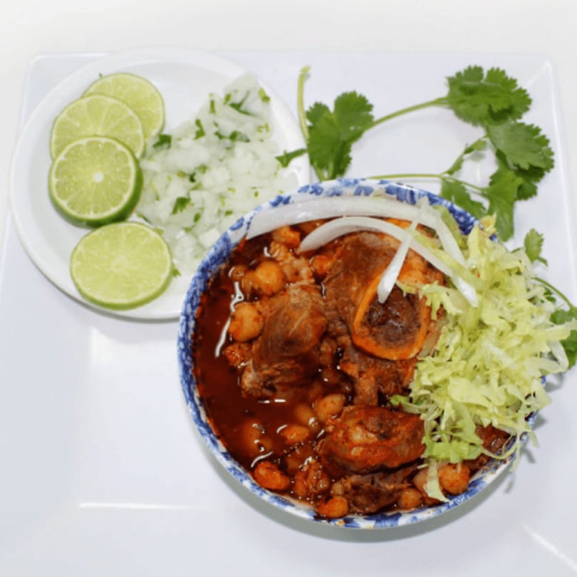 A bowl of pozole with meat, hominy, and red broth, garnished with shredded lettuce and onions.