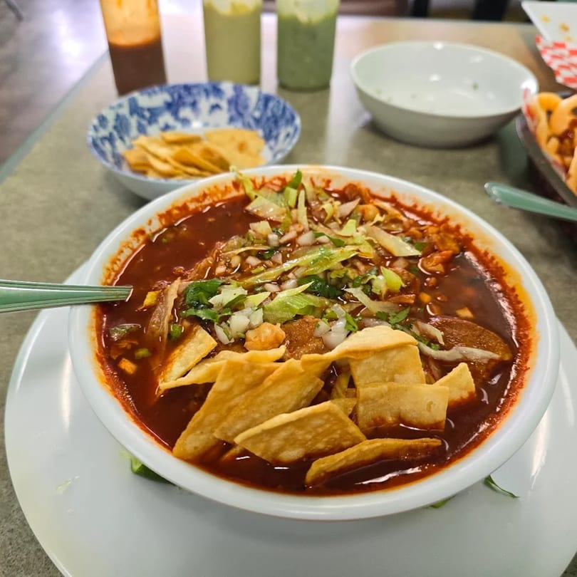A bowl of red soup with tortilla strips, lettuce, and onions, with sauces and chips in the background.