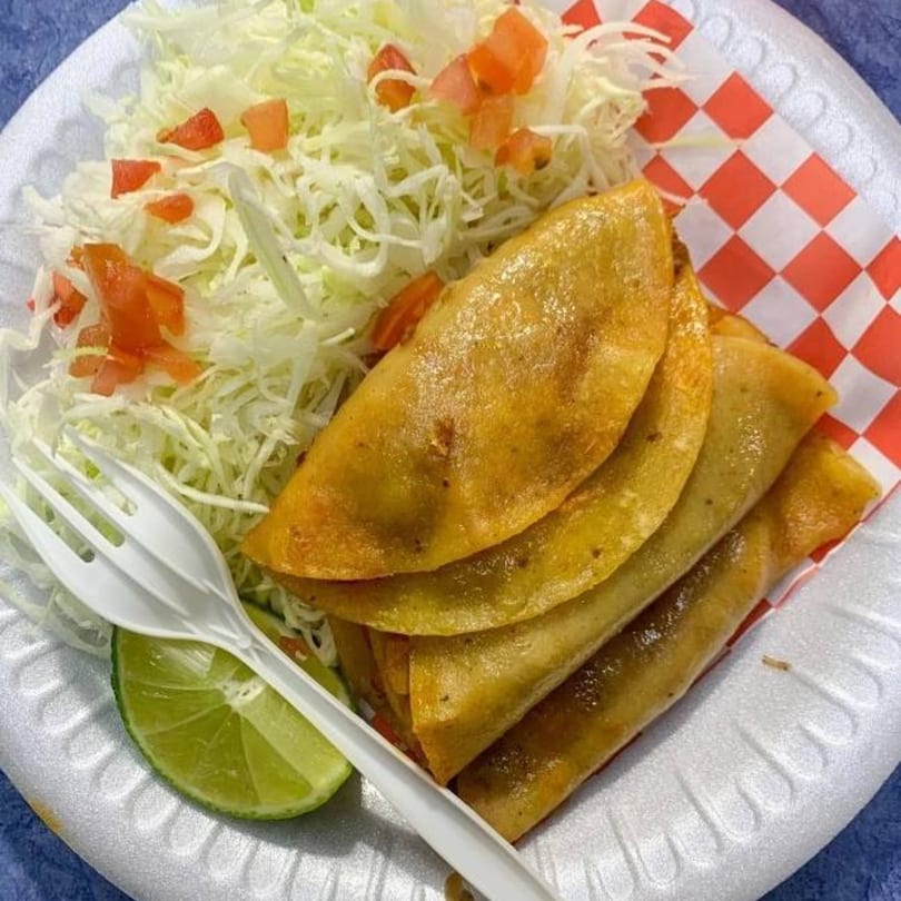 Four golden fried tacos with shredded lettuce, diced tomatoes, and a lime wedge on a white plate.