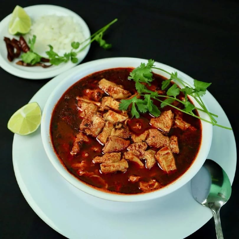 A bowl of menudo with cilantro, lime, and chopped onions on a white plate.
